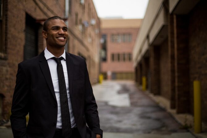 A man in a black suit and tie stands in an urban alleyway, smiling. Brick buildings line the alley on both sides.