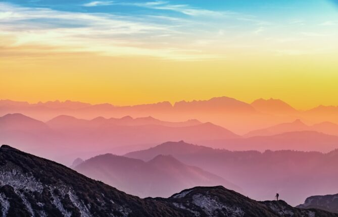 Silhouetted mountain range during sunrise with layers of hills fading into the horizon, sky transitioning from blue to orange.