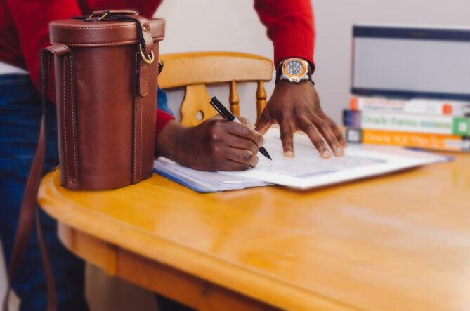 Person in a red shirt signing a document at a wooden table with a leather bag and stacked books nearby.