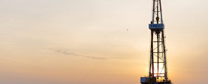 An oil drilling rig at sunset in an open field. A large truck and a container are visible to the left of the rig.